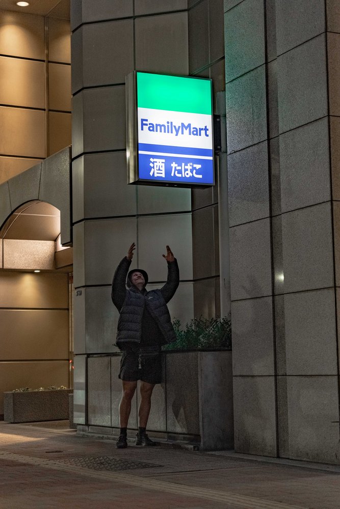 Hugh posing under a FamilyMart convenience store sign on a Tokyo street at night — Zen Den Studios