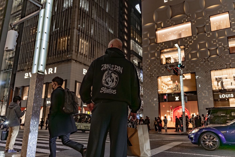 Hugh in a Zen Den Studios hoodie at a busy Ginza intersection at night with luxury stores in the background — Zen Den Studios