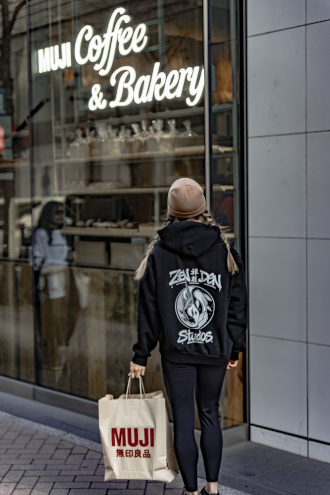 Liz from behind in a Zen Den Studios hoodie outside Muji Coffee and Bakery, holding a Muji tote bag — Zen Den Studios