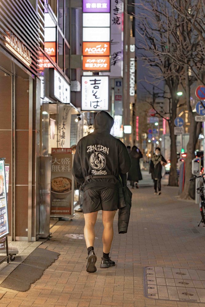Hugh from behind in a Zen Den Studios hoodie walking through a neon-lit Tokyo street at night — Zen Den Studios